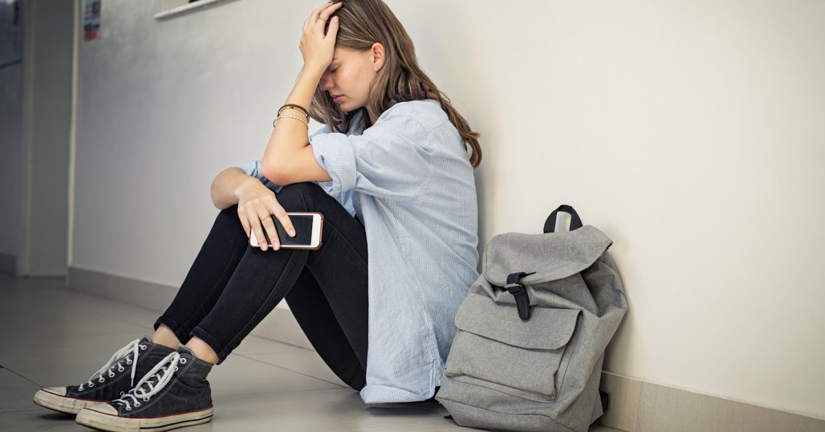 Upset and depressed girl holding smartphone sitting on college campus floor holding head. University sad student suffering from depression sitting on floor at high school. Lonely bullied teen in difficulty with copy space.