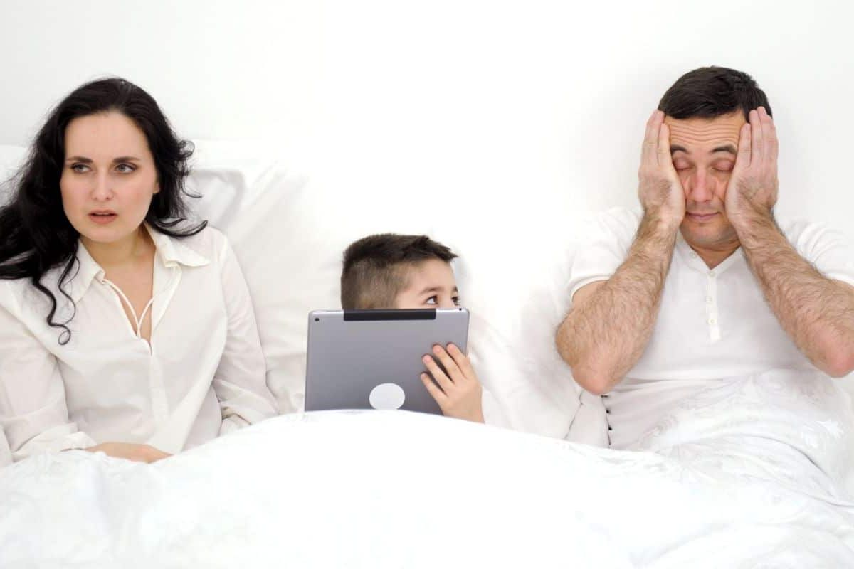 family in bed child on ipad parents look tired and miserable.