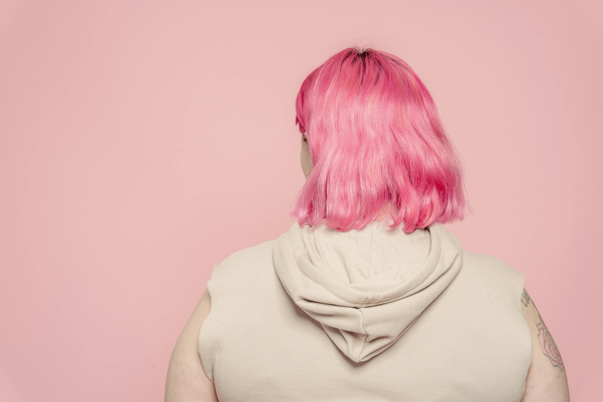 Back view of faceless female with dyed hair wearing sleeveless shirt standing against pink background