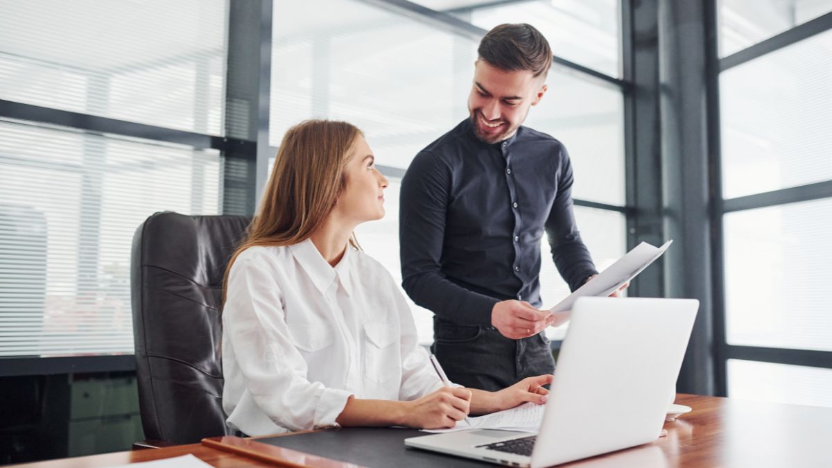 woman sitting at computer talking to man at work