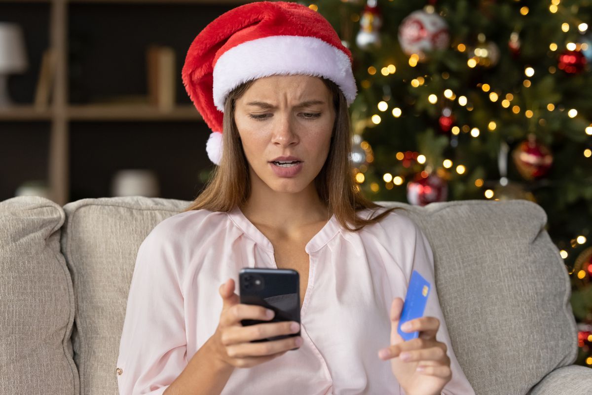 Woman in a santa hat looking at her phone holding a credit card upset.