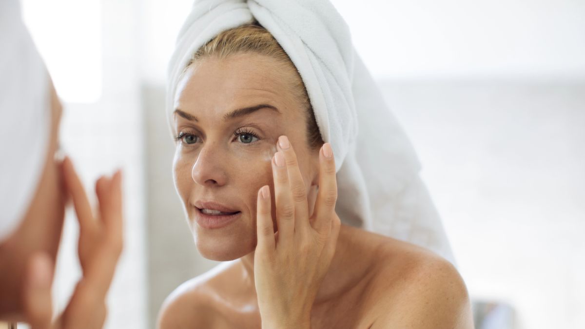 woman using skin cream on face in mirror