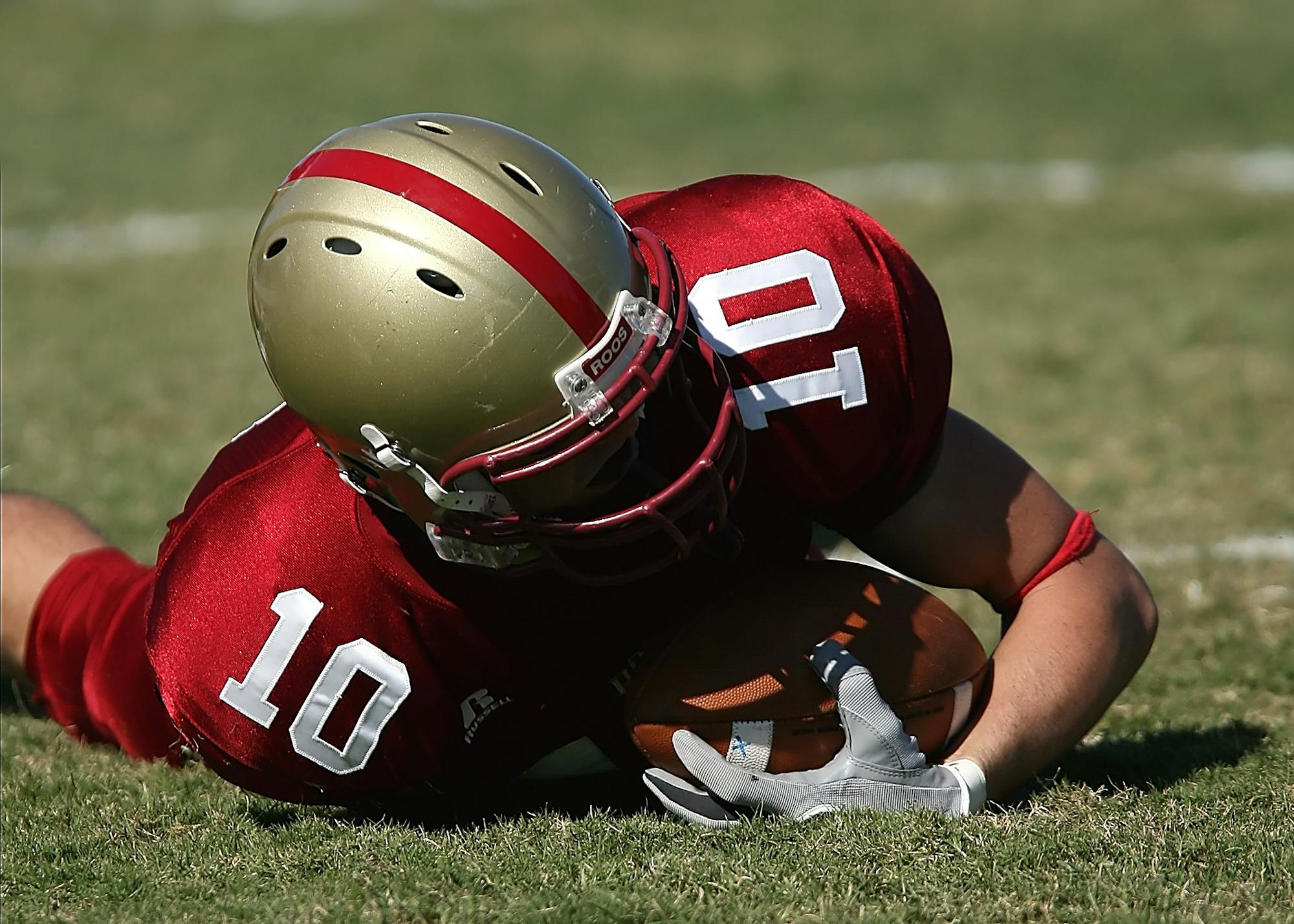 American football player wearing red uniform holding ball on grassy field during day game.