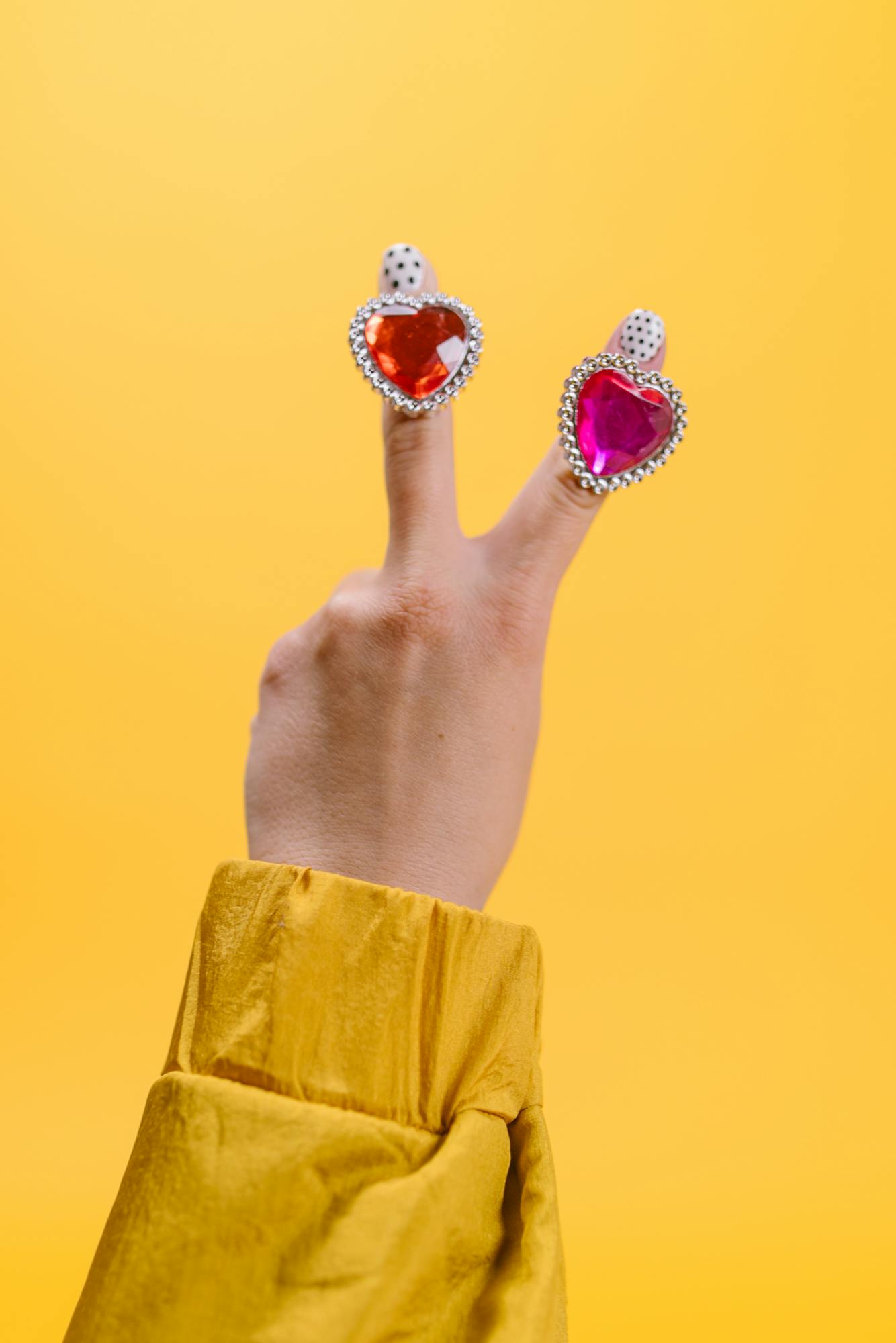 A hand making a peace sign with heart-shaped rings on a vibrant yellow background.