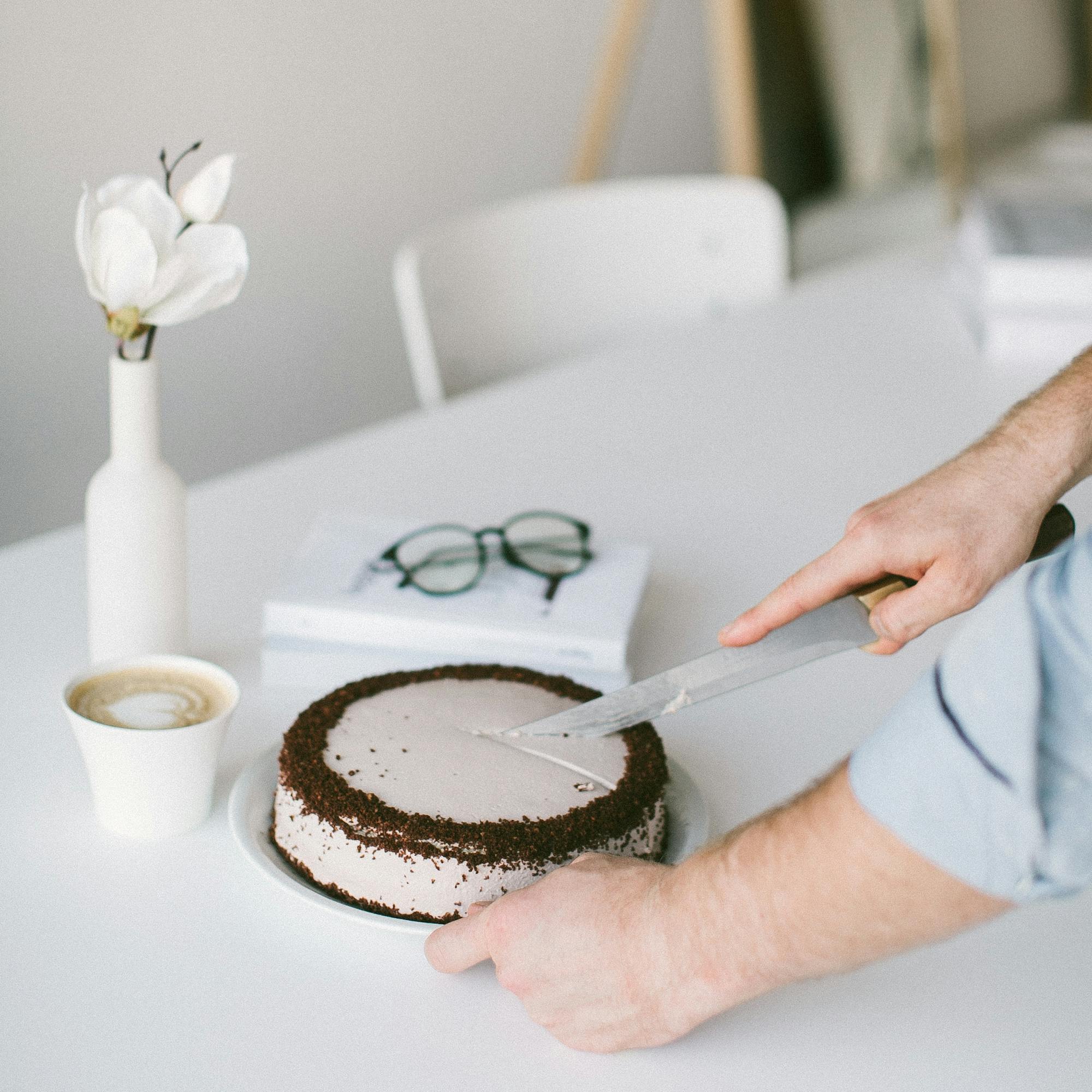 A person cutting a chocolate cake on a minimalist table with coffee and books. Ideal for lifestyle and food concepts.