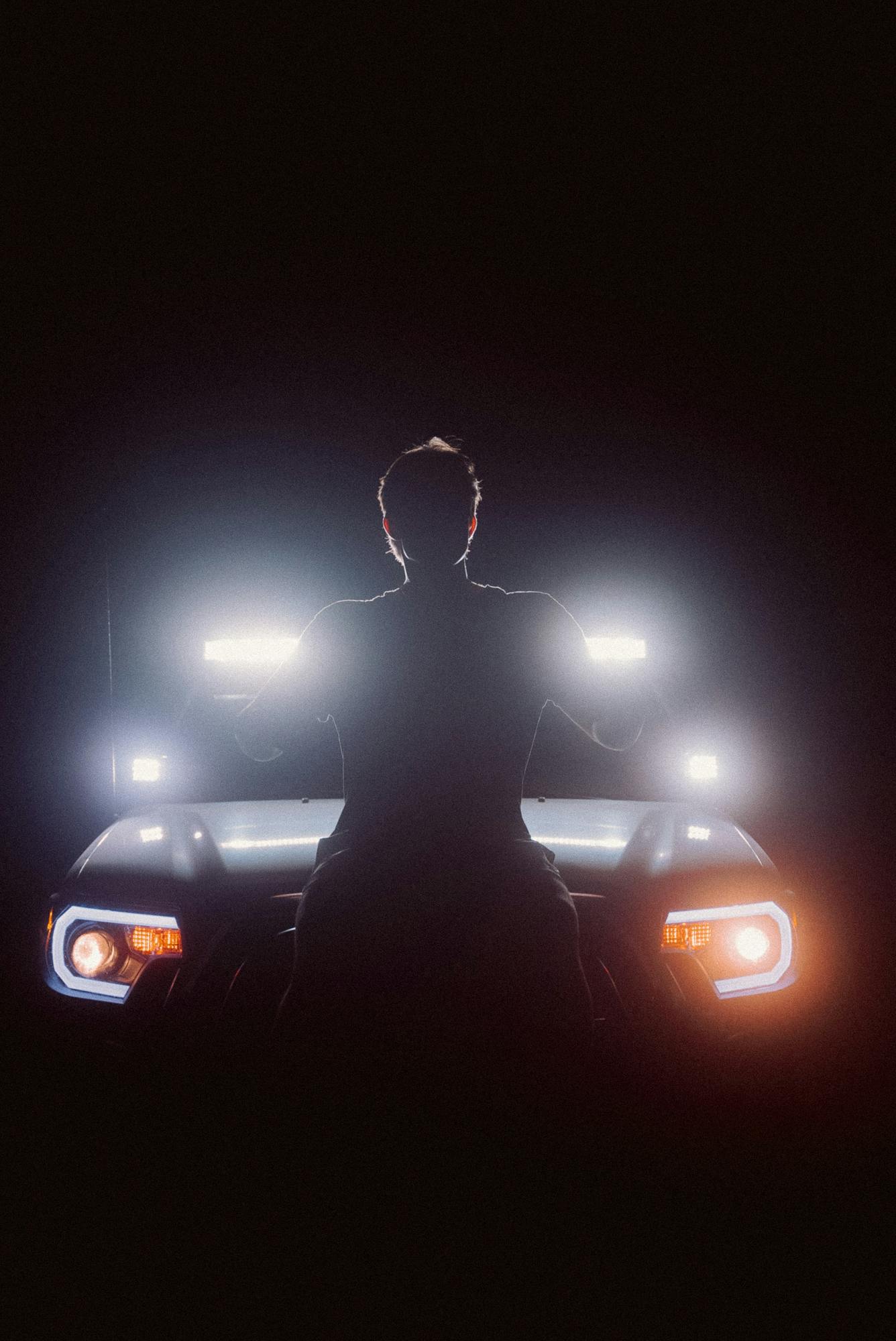 Silhouette of a man standing in front of a car with bright headlights at night, creating a dramatic scene.