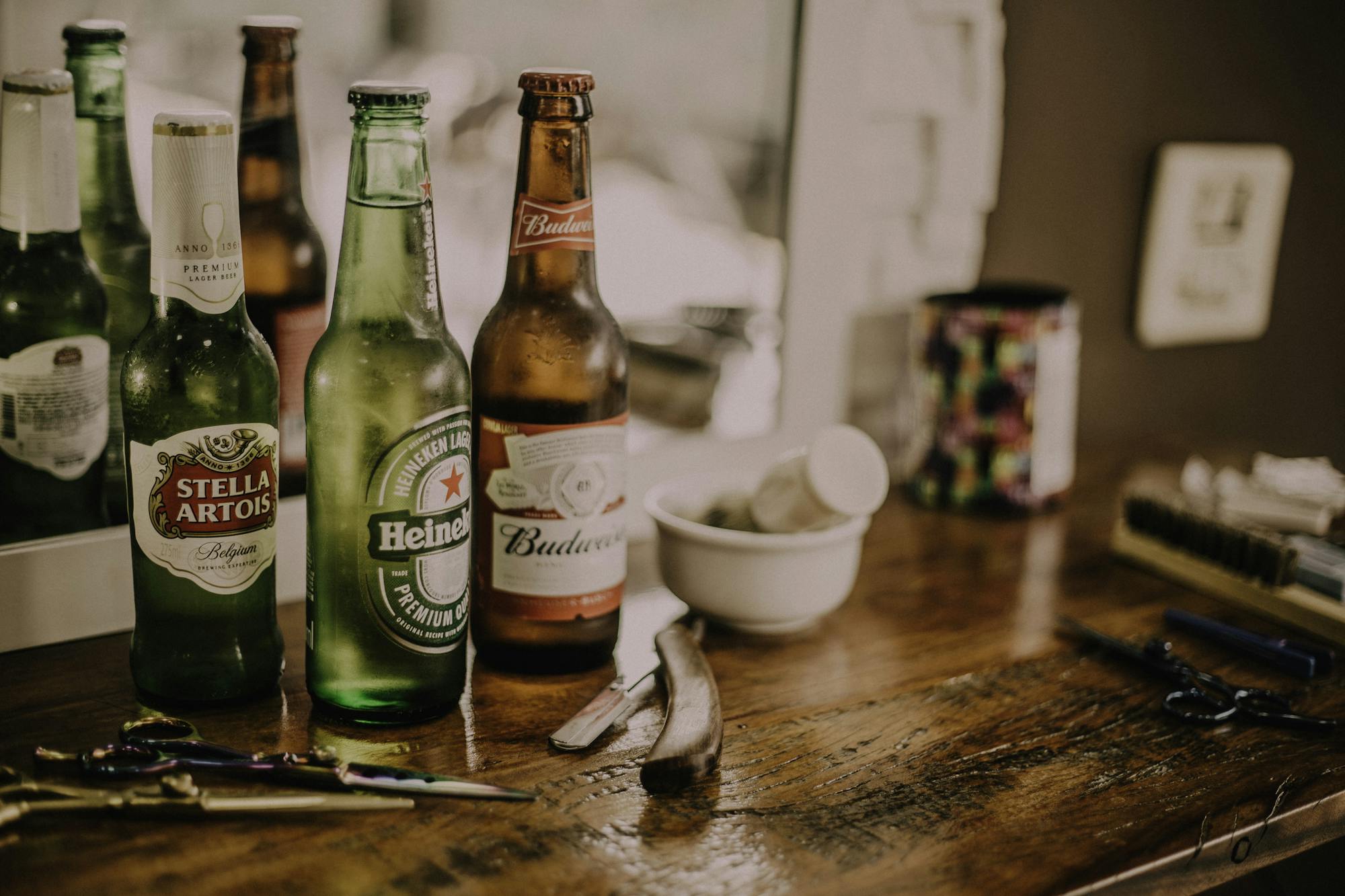 Beer bottles and barber tools on a wooden barbershop counter with a blurred mirror reflection.