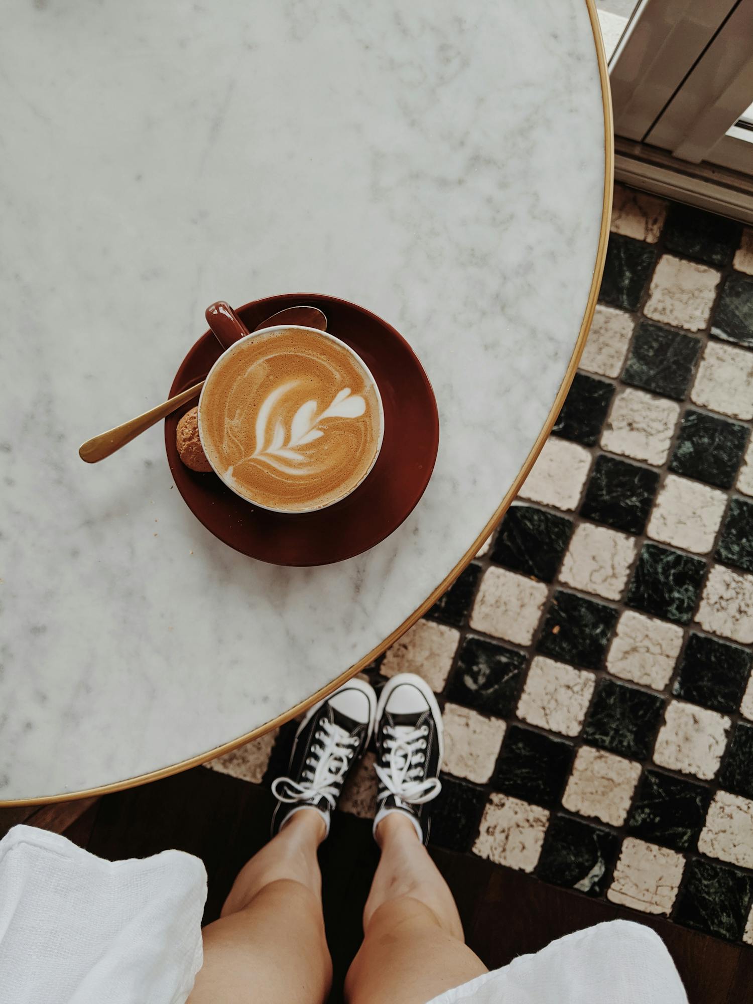 Overhead view of a latte with art on a marble table, sneakers visible on checkered floor.